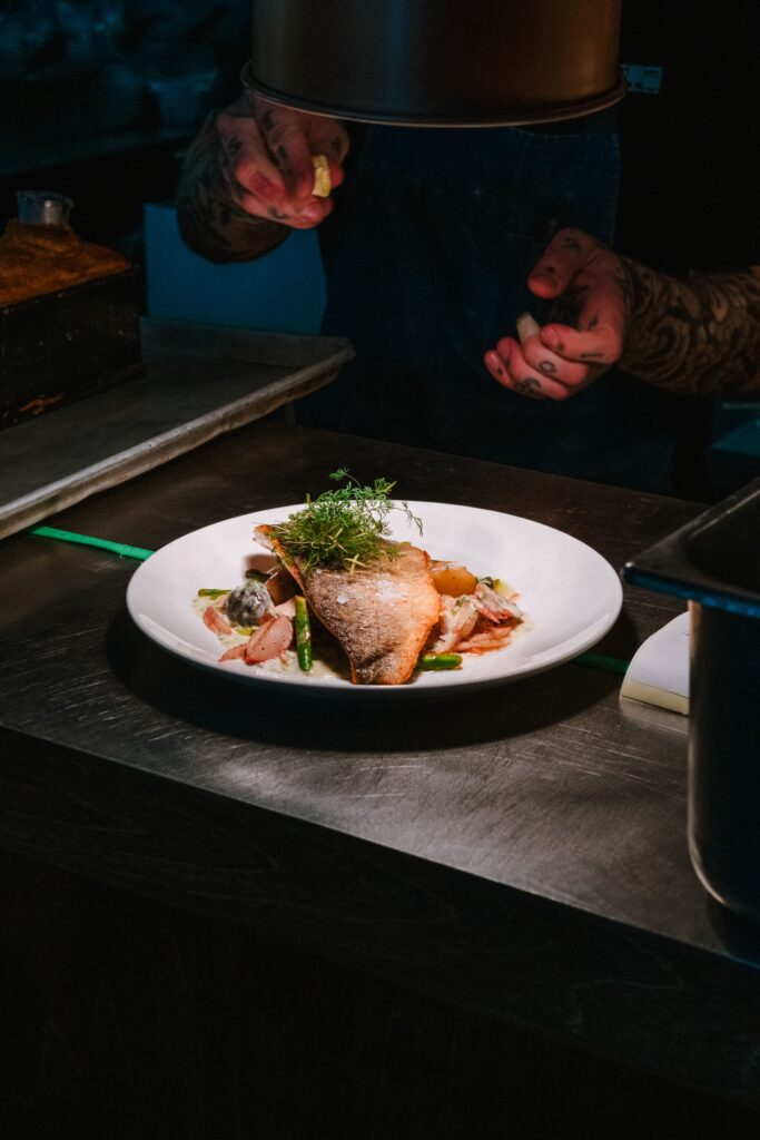 A chef carefully finishes a plated dish under the warm light of a professional kitchen. The composition highlights a beautifully prepared piece of fish, seasonal vegetables and delicate herbs, capturing the precision, craftsmanship and attention to detail behind a refined dining experience.