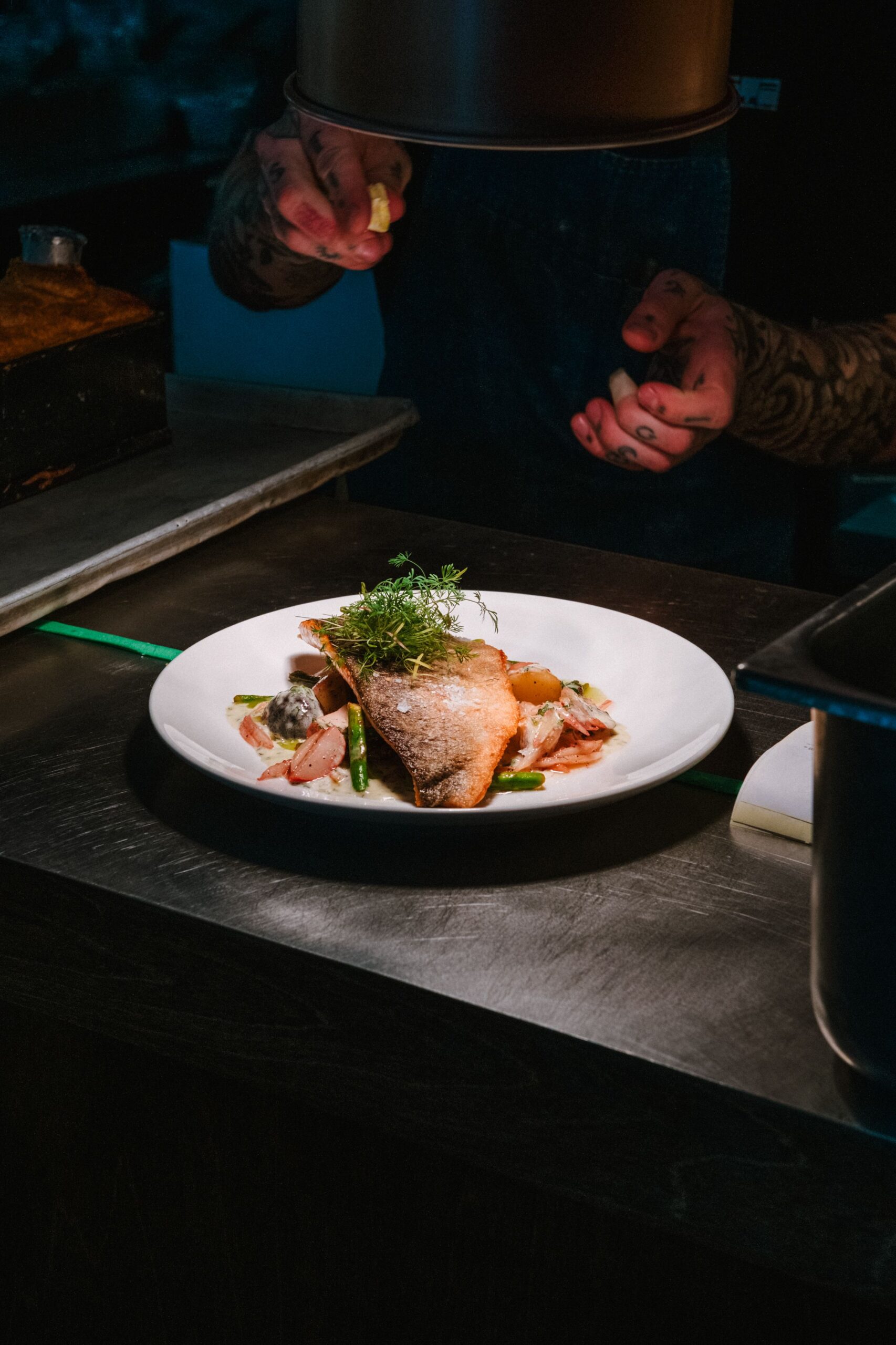 A chef carefully finishes a plated dish under the warm light of a professional kitchen. The composition highlights a beautifully prepared piece of fish, seasonal vegetables and delicate herbs, capturing the precision, craftsmanship and attention to detail behind a refined dining experience.