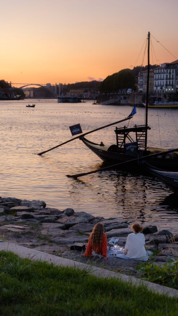 A peaceful sunset over the Douro River in Porto, with traditional boats resting on the water and the Dom Luís I Bridge visible in the distance. In the foreground, two people sit by the river enjoying a quiet picnic, while the warm evening light reflects across the water and the historic riverside buildings create an intimate, timeless atmosphere.